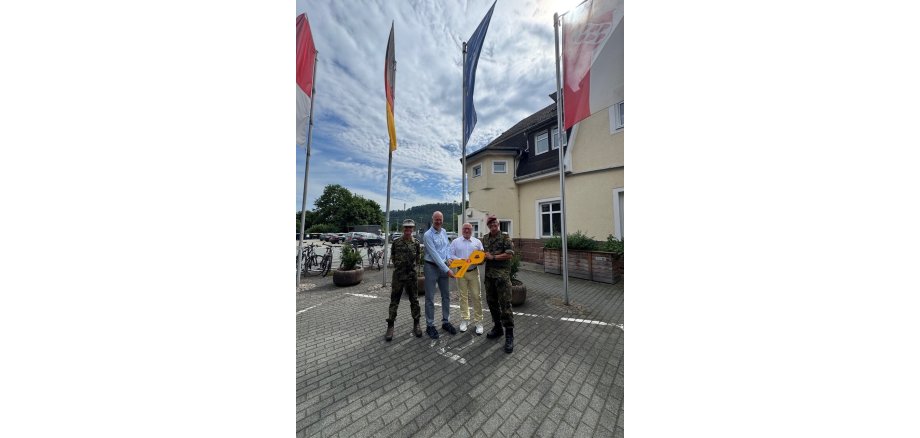 Oberst Döring und Hauptmann Würz übergeben die Gelbe Schleife an den Bürgermeister Dr. Ried und den Vorsitzenden der Gemeindevertretung vor dem Rathaus; die hessische, deutsche, europäische und Cölber Flaggen sind gehisst im Hintergrund zu sehen