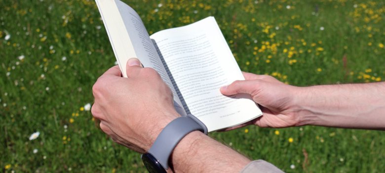 Zwei Hände halten ein aufgeklapptes Buch. Im Hintergrund sieht man eine grüne Wiese.